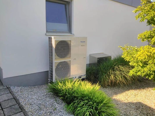 Außen an einem modernen Einfamilienhaus steht eine Wärmepumpe im Garten zwischen grünen Pflanzen.